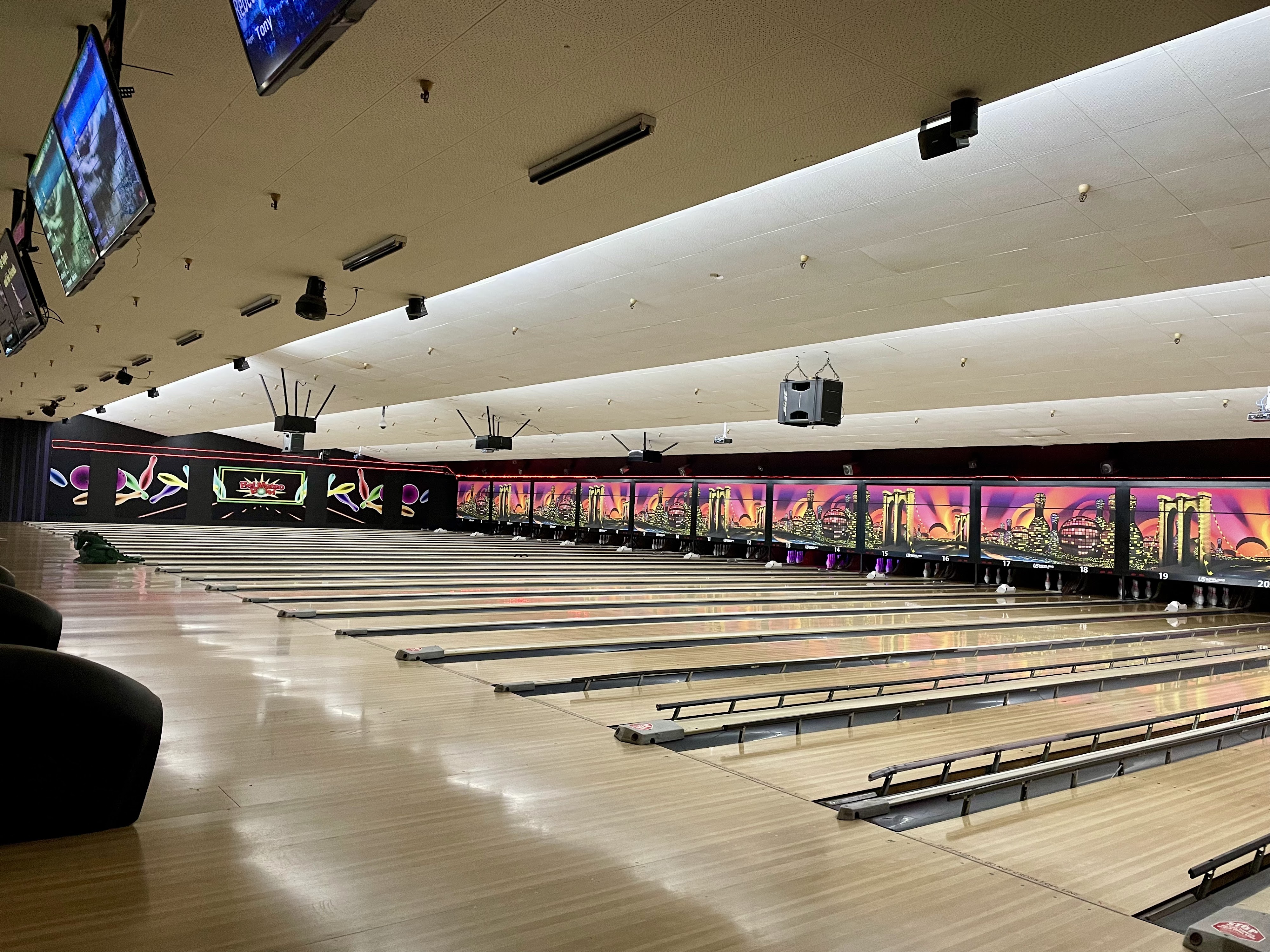 A wide view of many polished wooden bowling lanes with colorful city murals and the Bel Mateo Bowl logo.