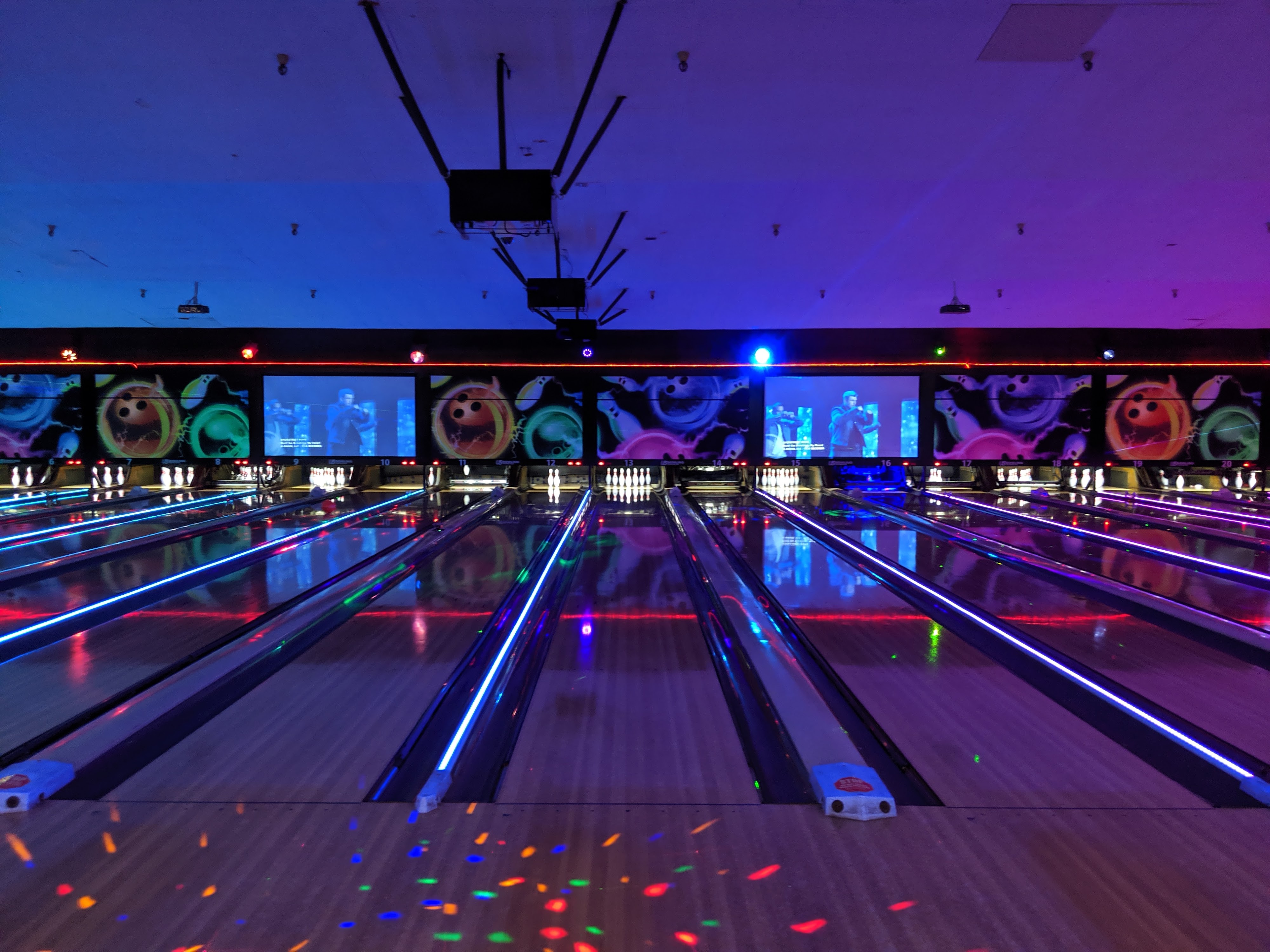 Symmetrical view of bowling lanes under vibrant purple and blue cosmic lighting with video screens above the pins.