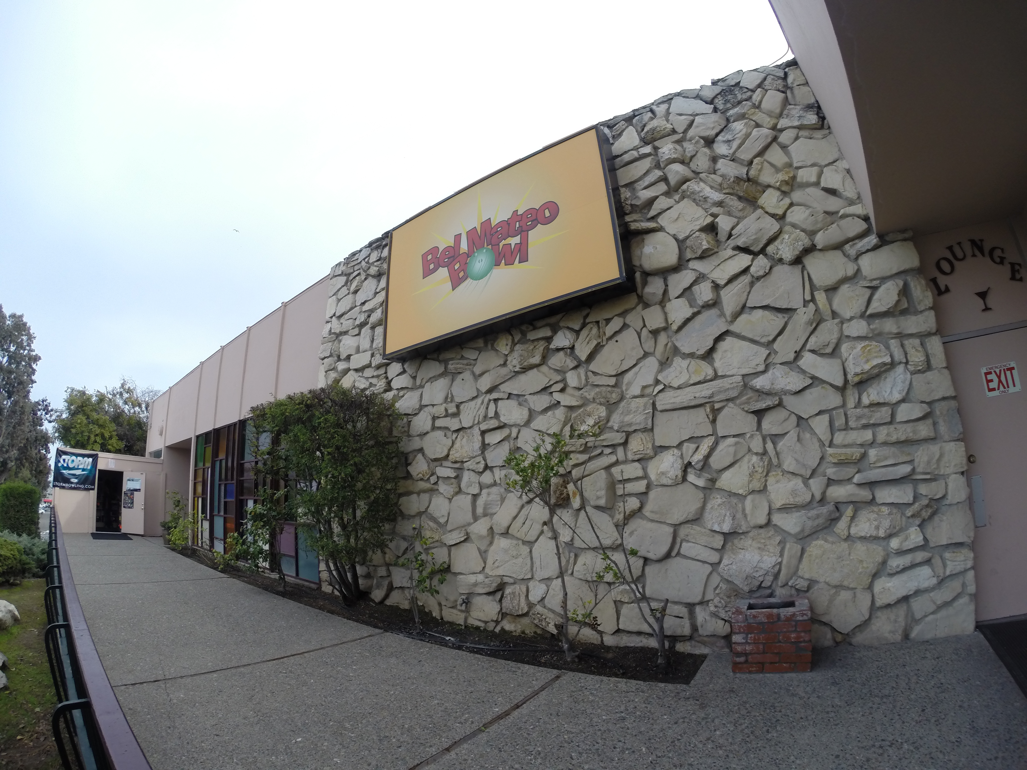 The exterior stone wall and logo sign of Bel Mateo Bowl under an overcast sky.