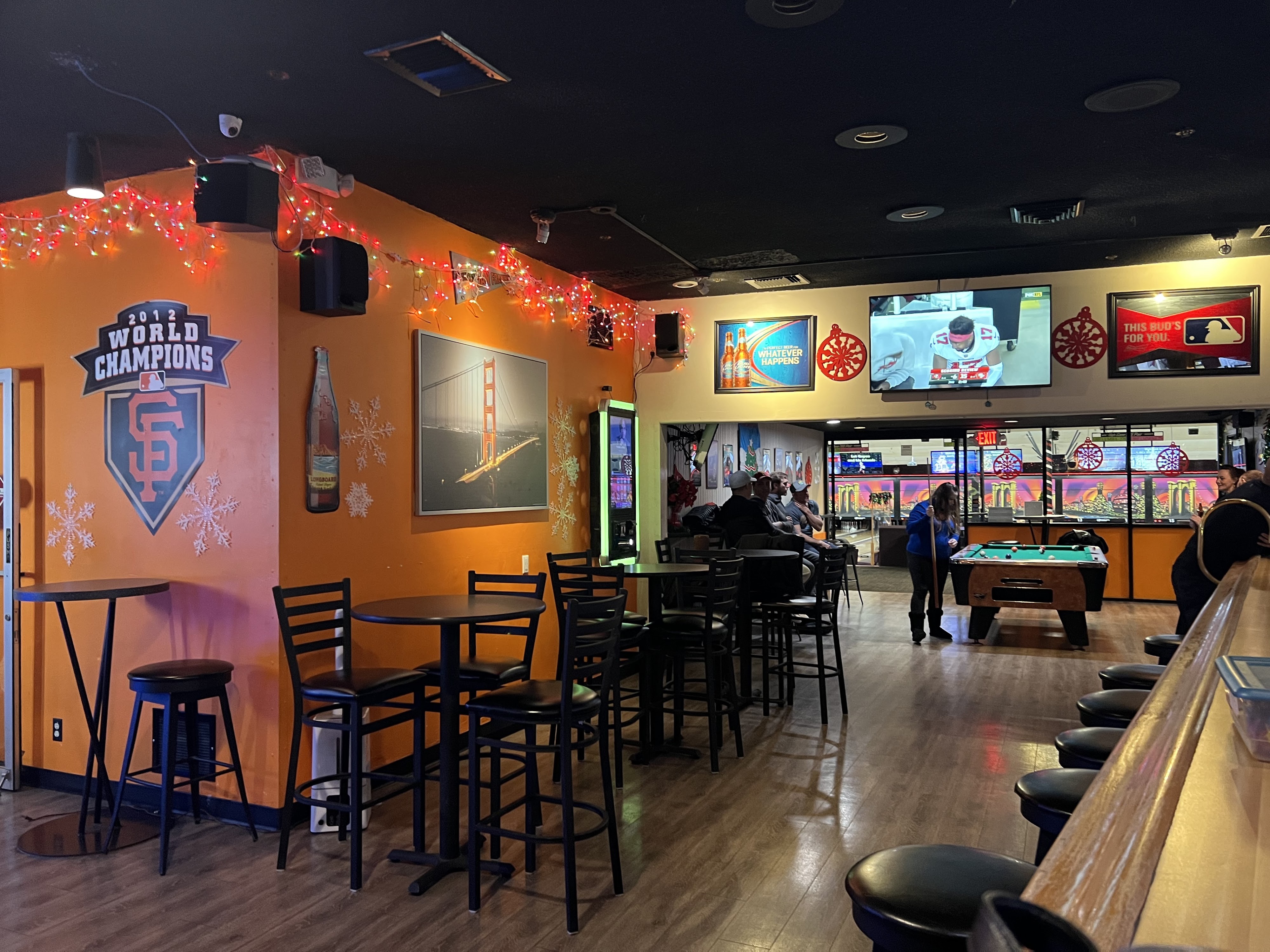 A sports bar lounge with orange walls, high-top tables, televisions showing sports, and a pool table.