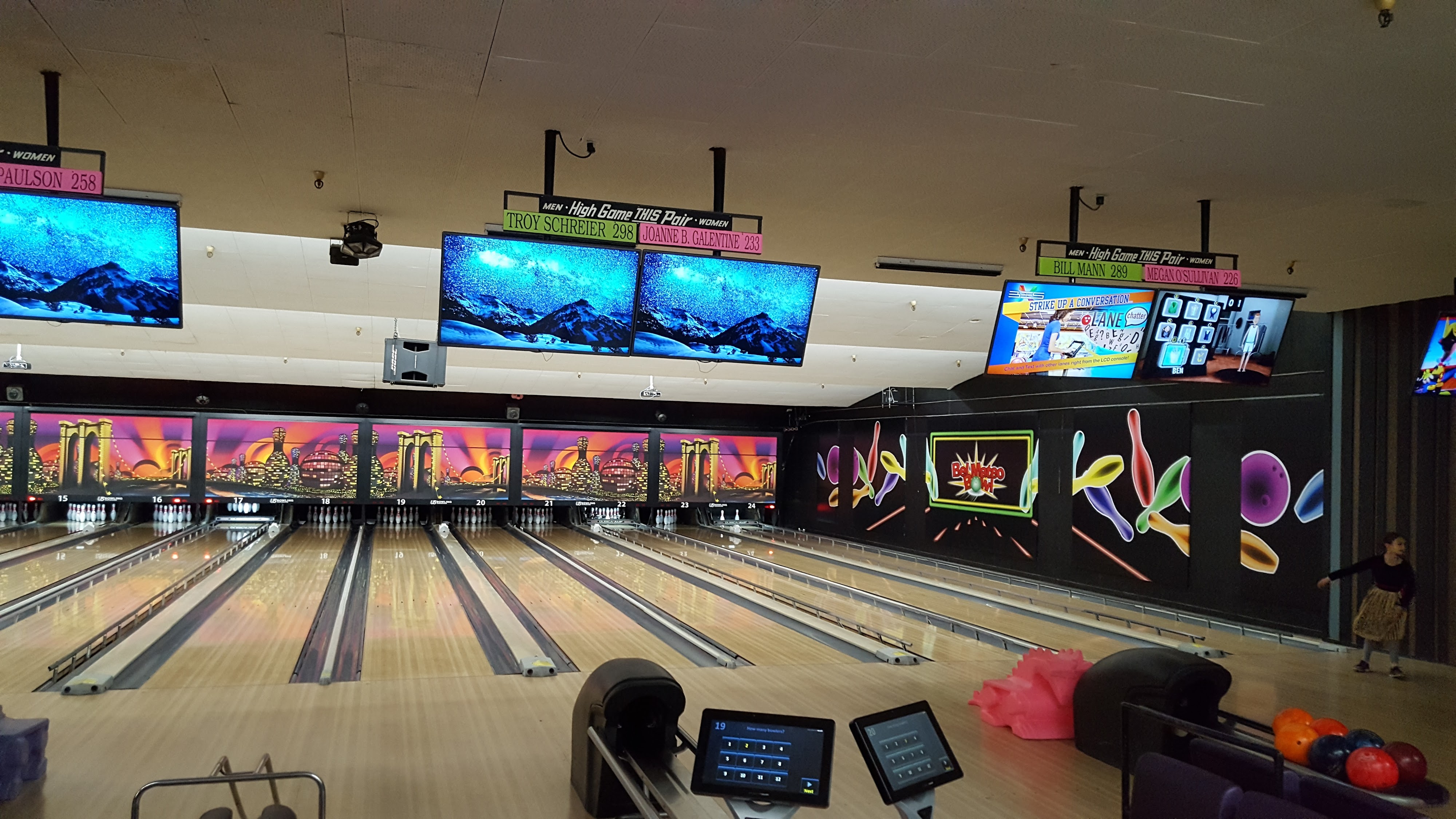 A wide view of several bowling lanes with digital scoring monitors and colorful city skyline murals above the pins.