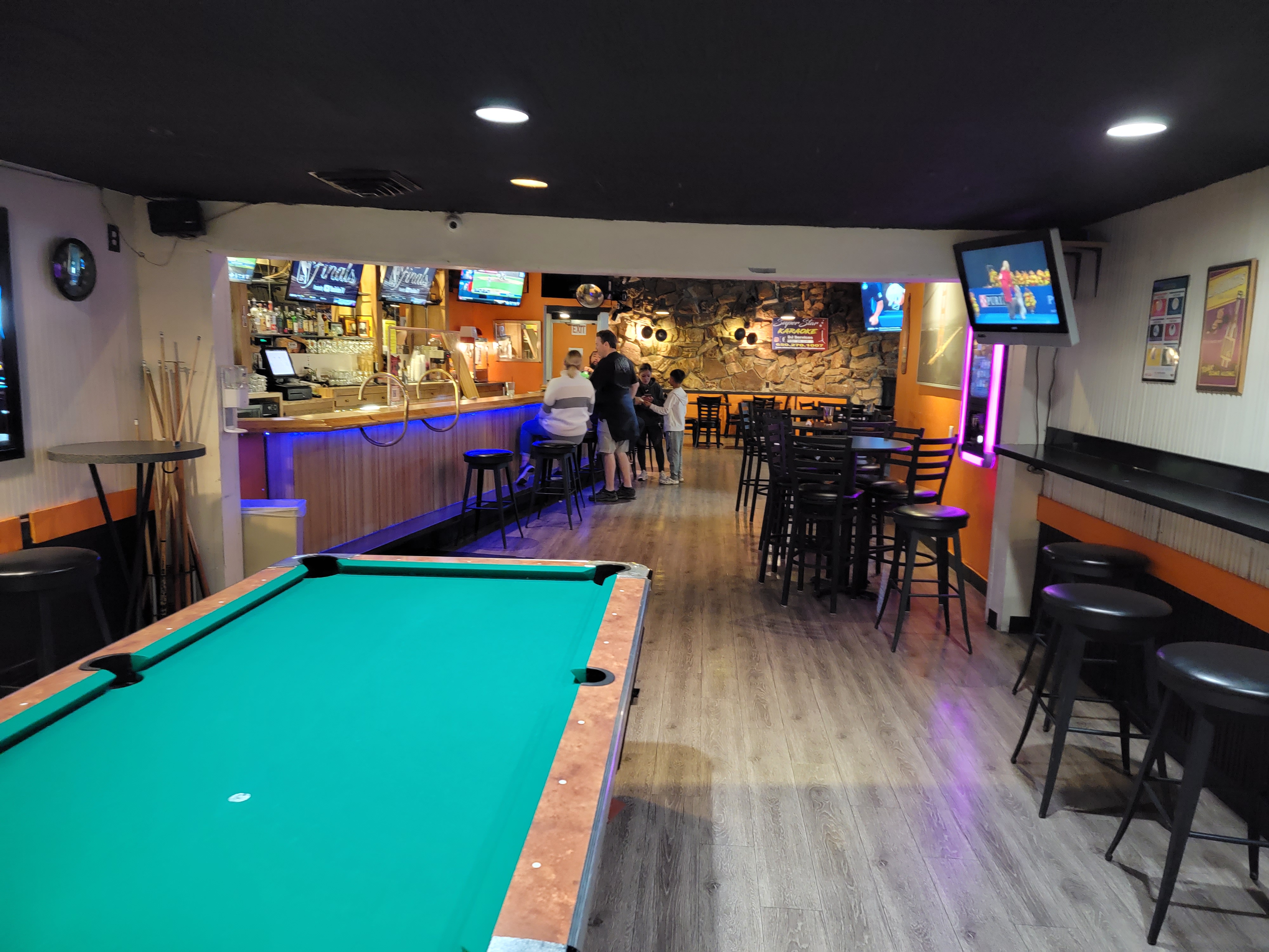 A green pool table in a spacious lounge area with a bar and television screens in the background.
