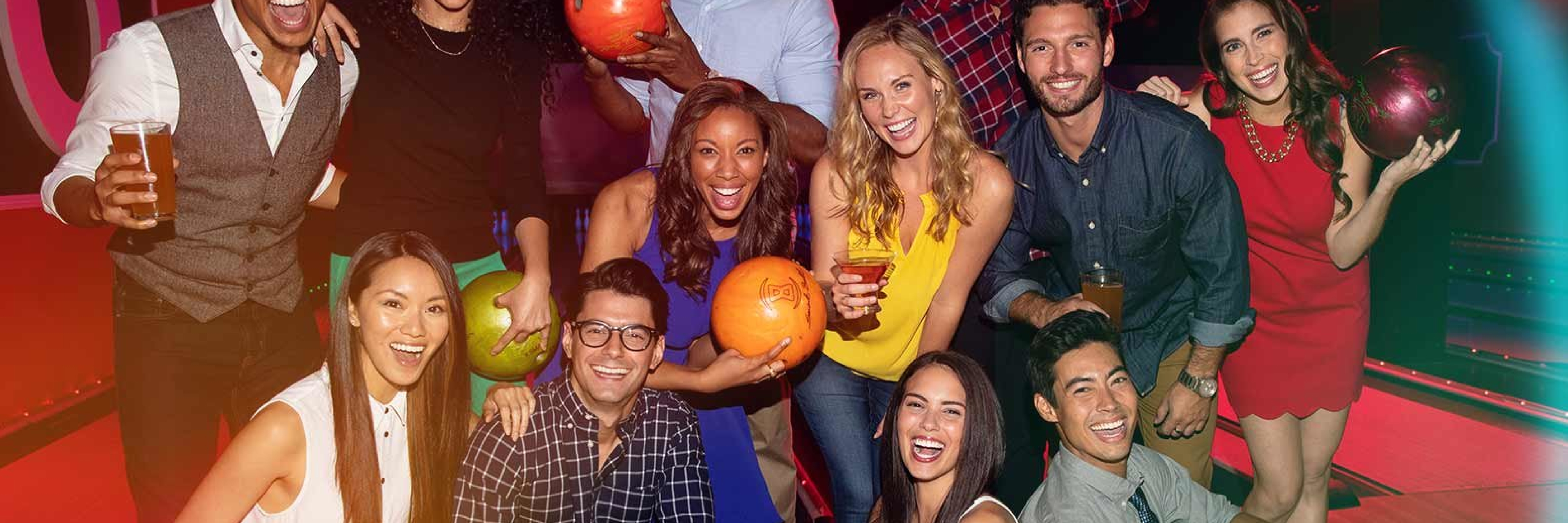 A large group of smiling friends holding bowling balls and drinks under neon lights at a bowling center.