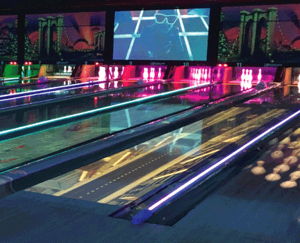 Bowling lanes glowing with neon blue and purple lights under large video screens in a dark alley.