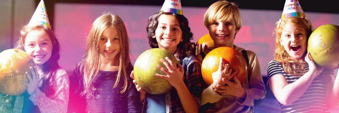 Five smiling children holding bowling balls and wearing birthday hats in a bowling alley with pink cosmic lighting.