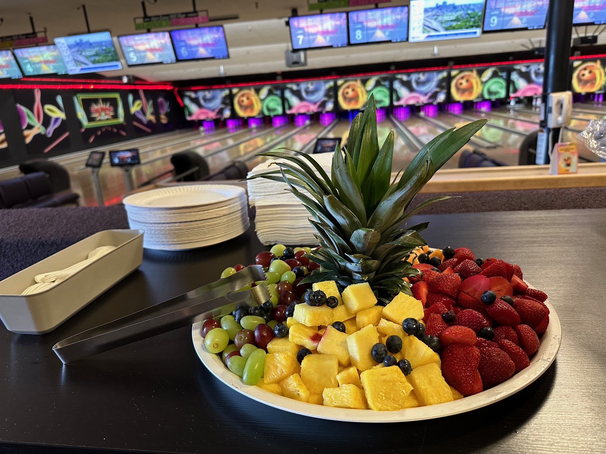 A fresh fruit platter with a pineapple crown in front of purple-lit bowling lanes.