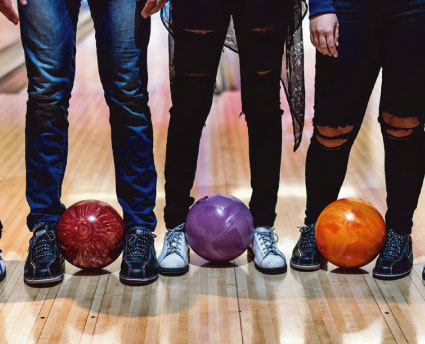 Three people standing on a bowling lane with red, purple, and orange bowling balls at their feet.
