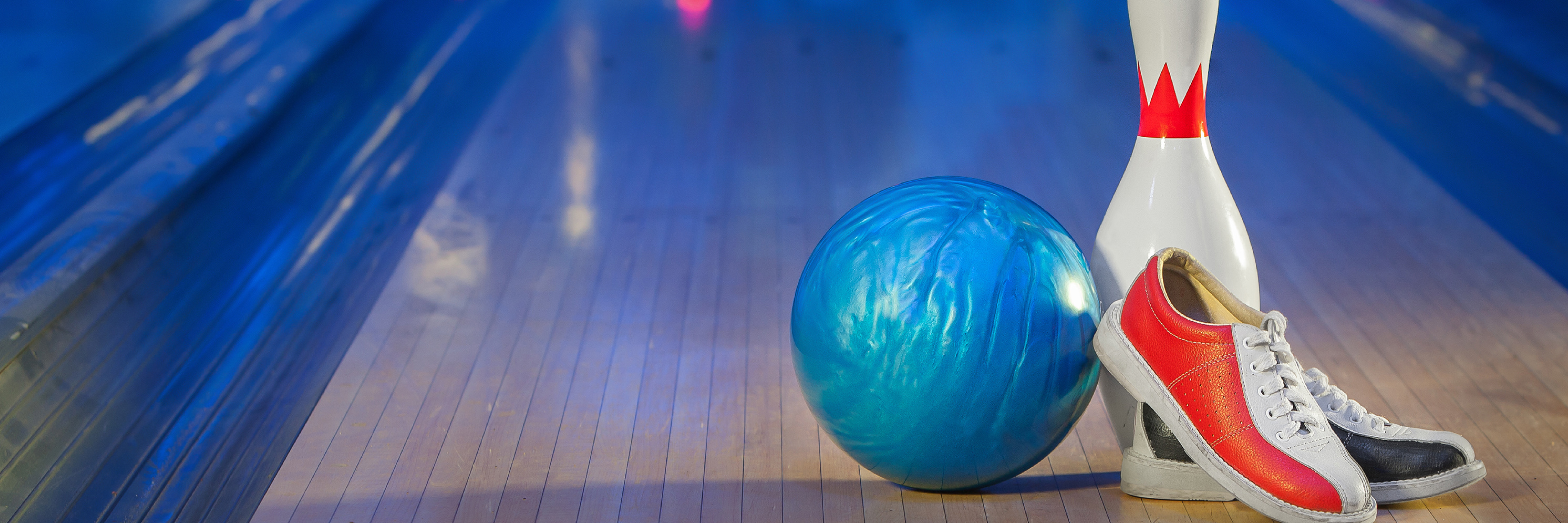 A blue bowling ball, a white bowling pin, and rental shoes sit on a wooden lane under atmospheric blue light.
