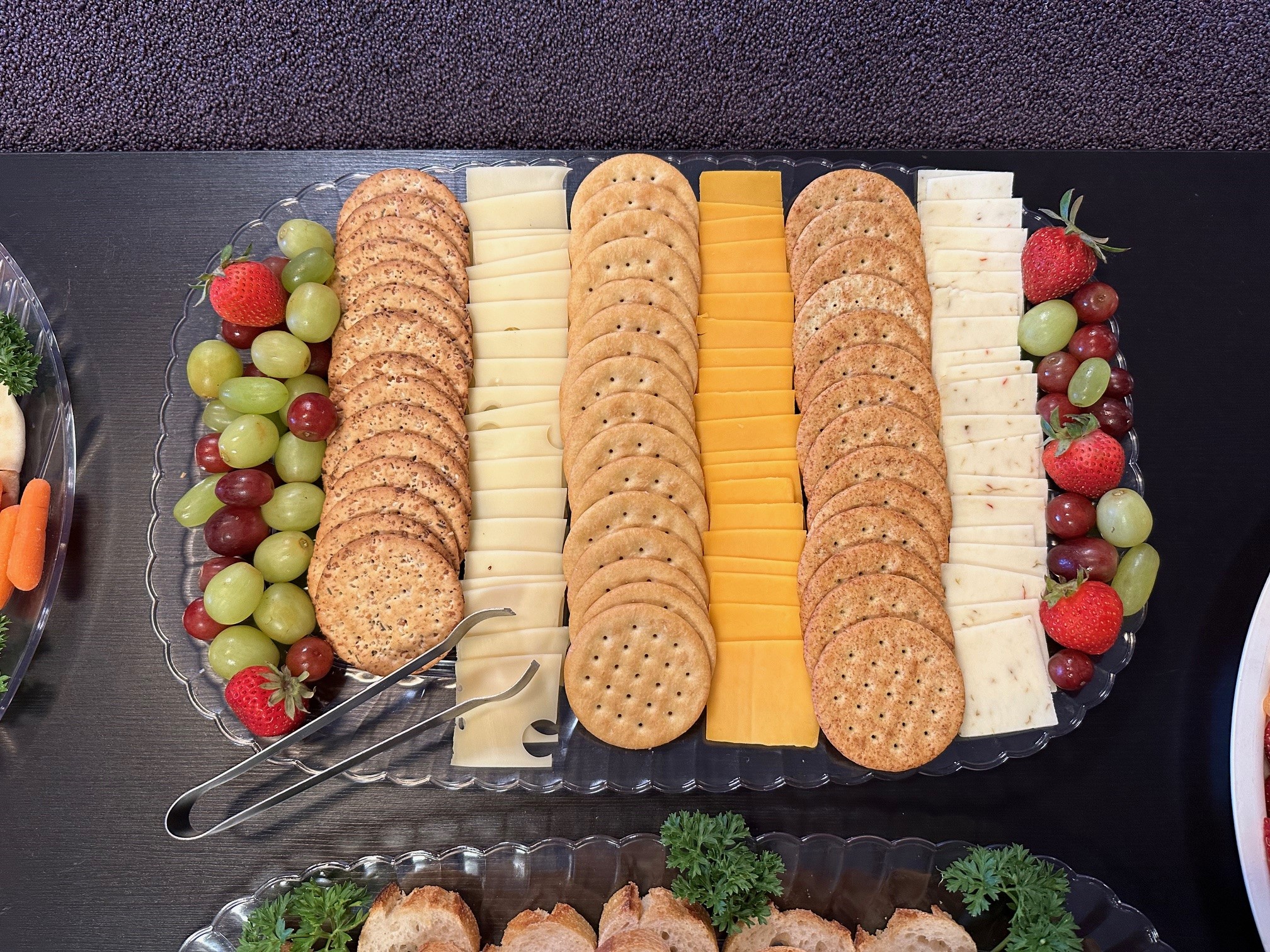 A top-down view of a catered platter featuring crackers, assorted cheeses, grapes, and strawberries.