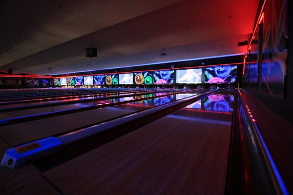 Polished bowling lanes under vibrant blue and red cosmic lighting with colorful video screens above the pins.