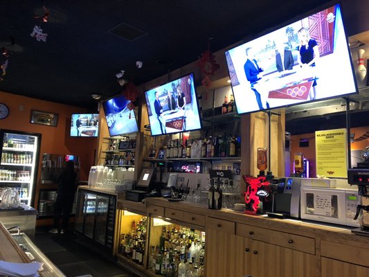 The full-service bar area at Bel Mateo Bowl featuring multiple TVs, wooden shelving, and various beverage selections.