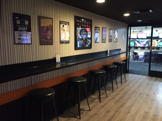 A row of black bar stools tucked under a long counter with framed posters on the wall and bowling lanes in the distance.