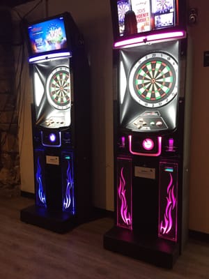 Two electronic dartboards with glowing blue and pink neon lights standing in a game room.