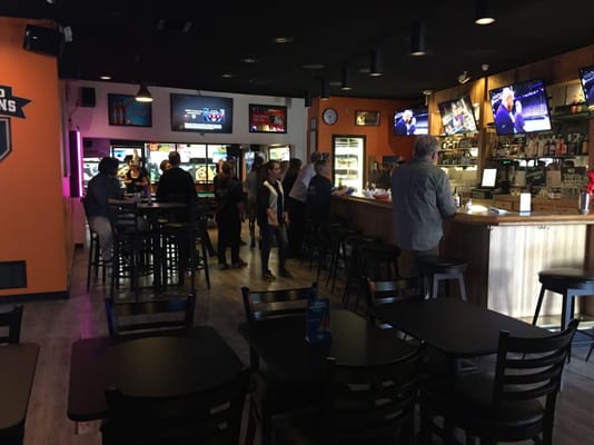 A dimly lit lounge and bar area with patrons sitting at the bar and multiple TVs on the wall.