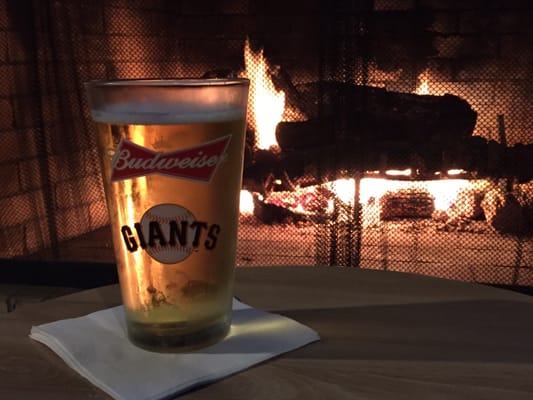 A pint of beer in a Giants logo glass sits on a table in front of a warm brick fireplace.