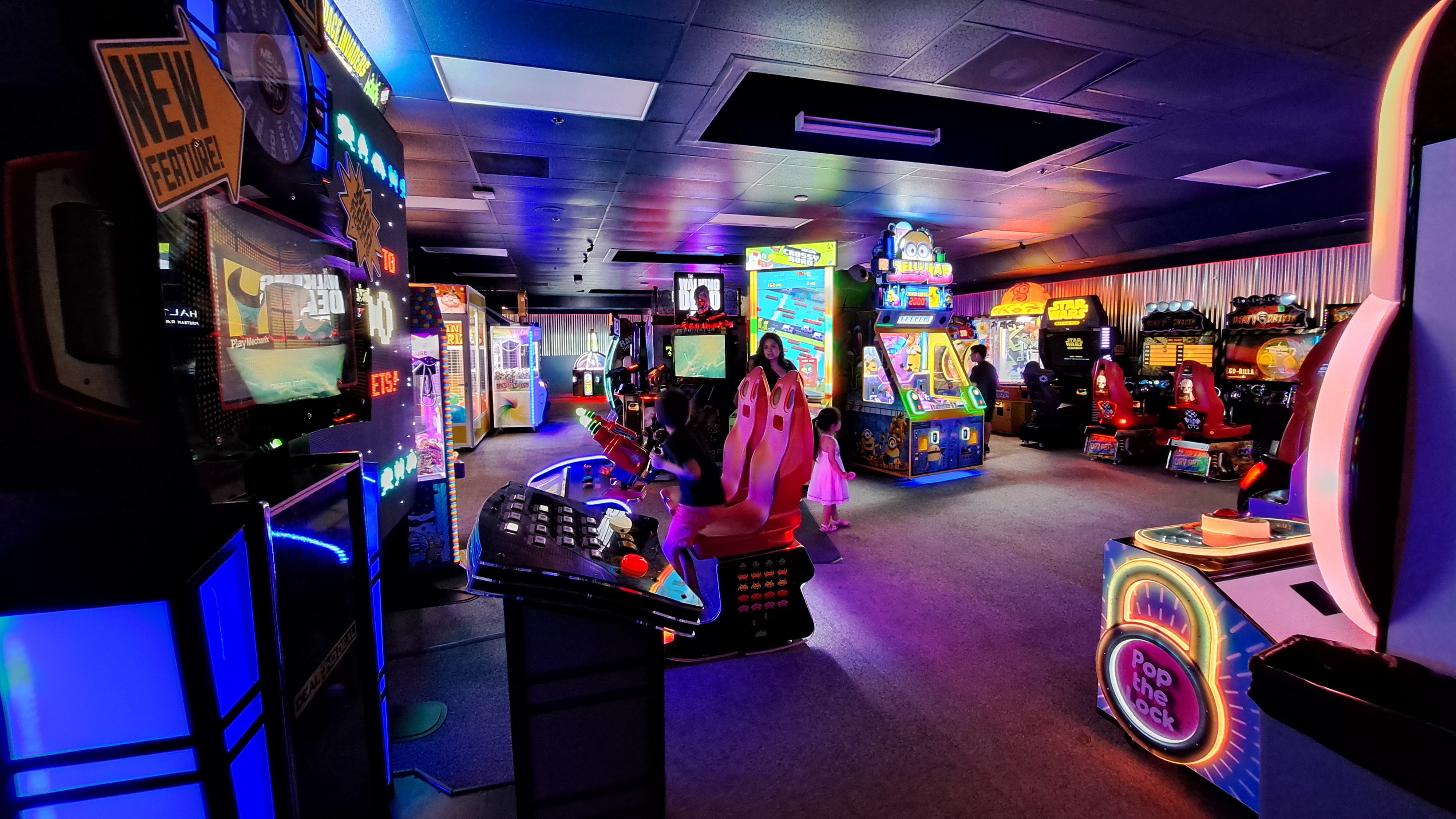 A wide view of a colorful arcade room filled with glowing video games and neon lights with a few people playing.