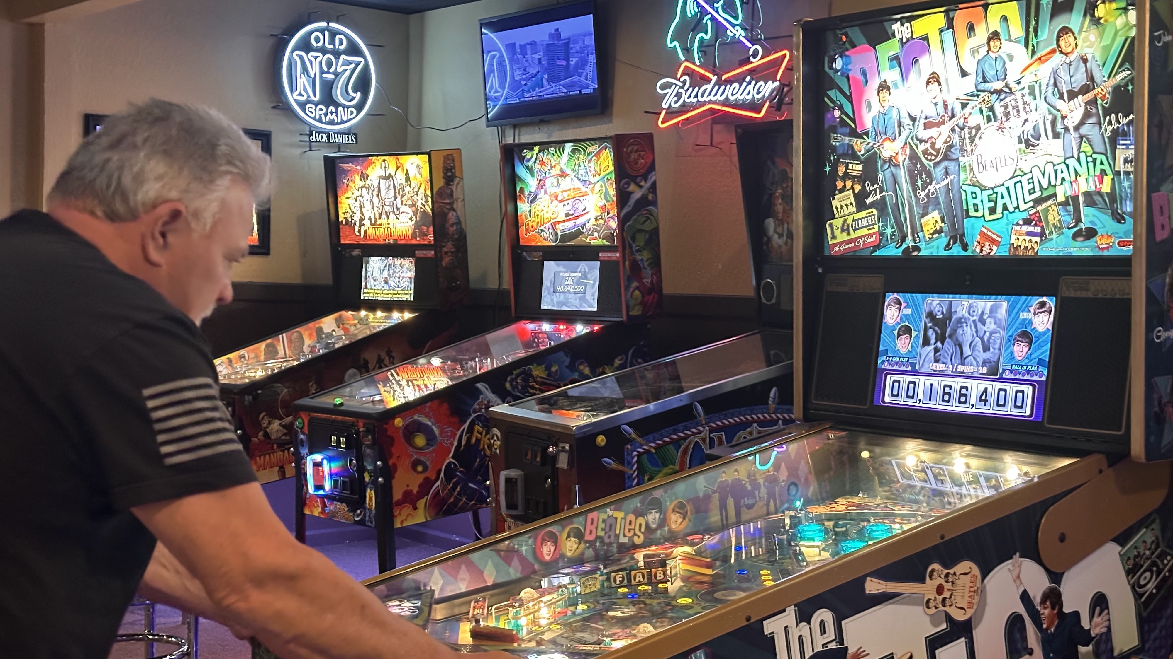 A man plays a Beatles-themed pinball machine in a dimly lit arcade with neon signs in the background.