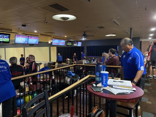League bowlers in matching shirts gather around tables and scoring monitors in a bowling center concourse.