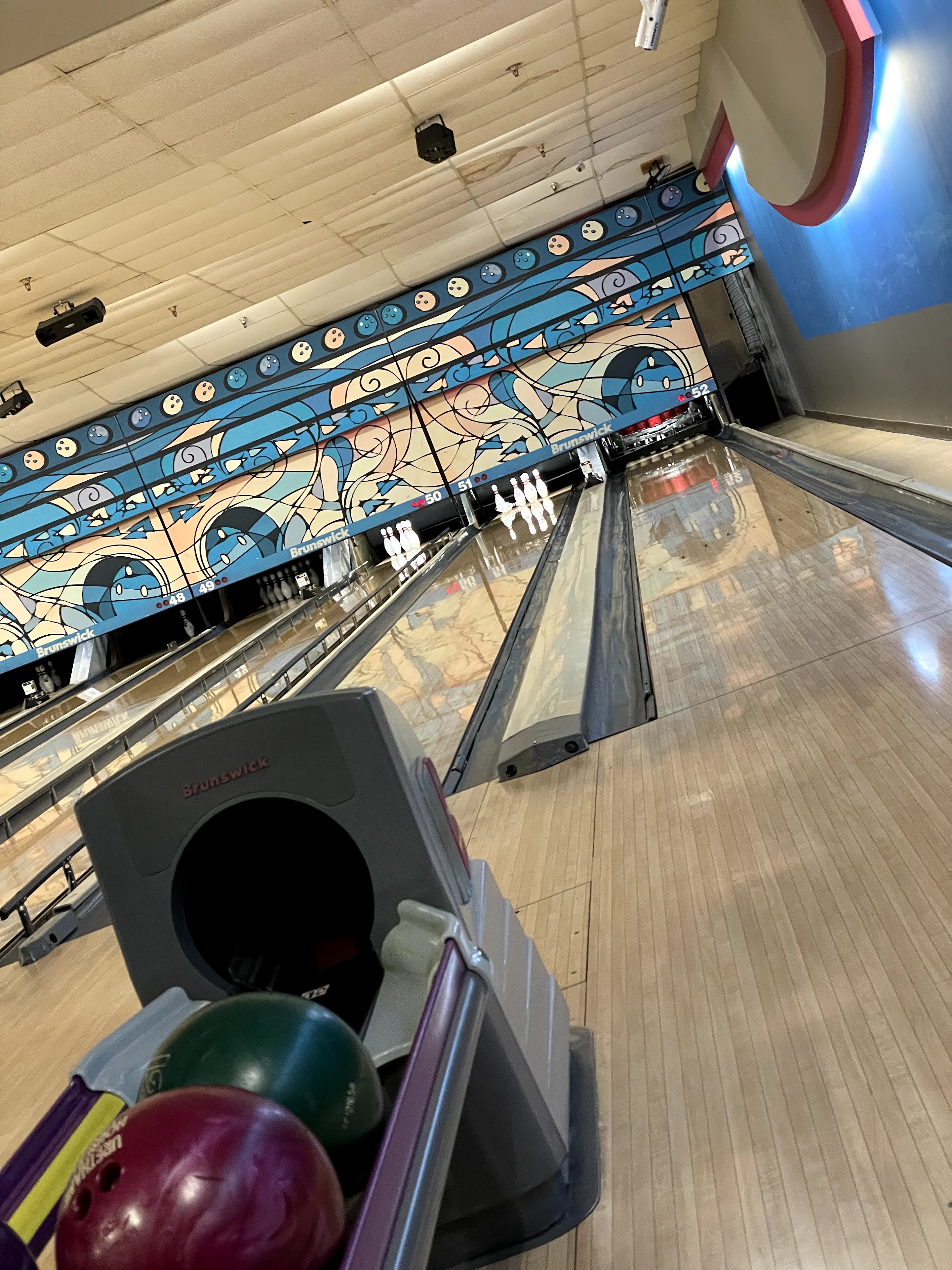 A tilted view of a bowling lane and ball return with colorful mural graphics on the back wall.