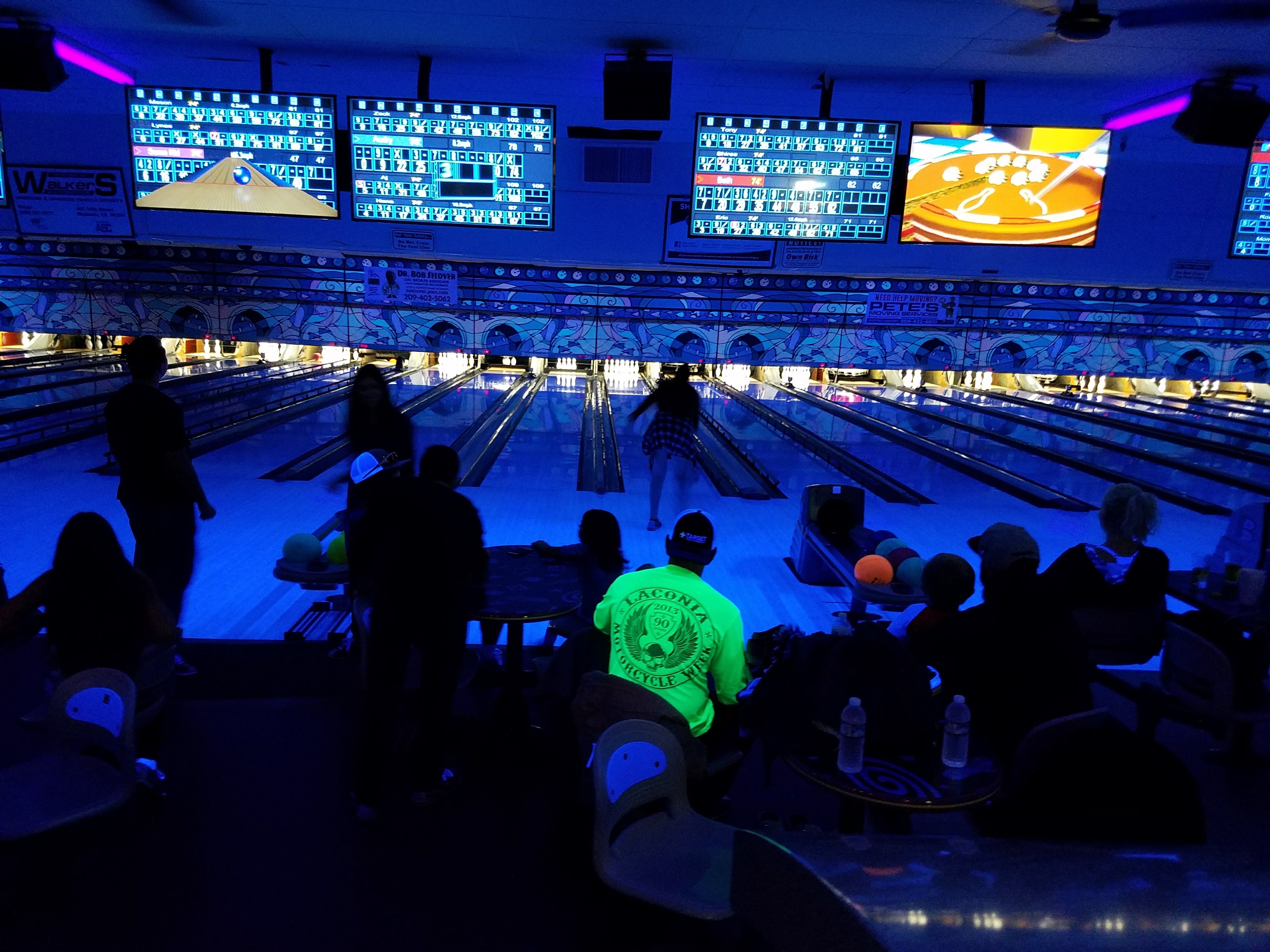 People bowling in a dimly lit center with vibrant blue glow lighting and digital scoring screens.