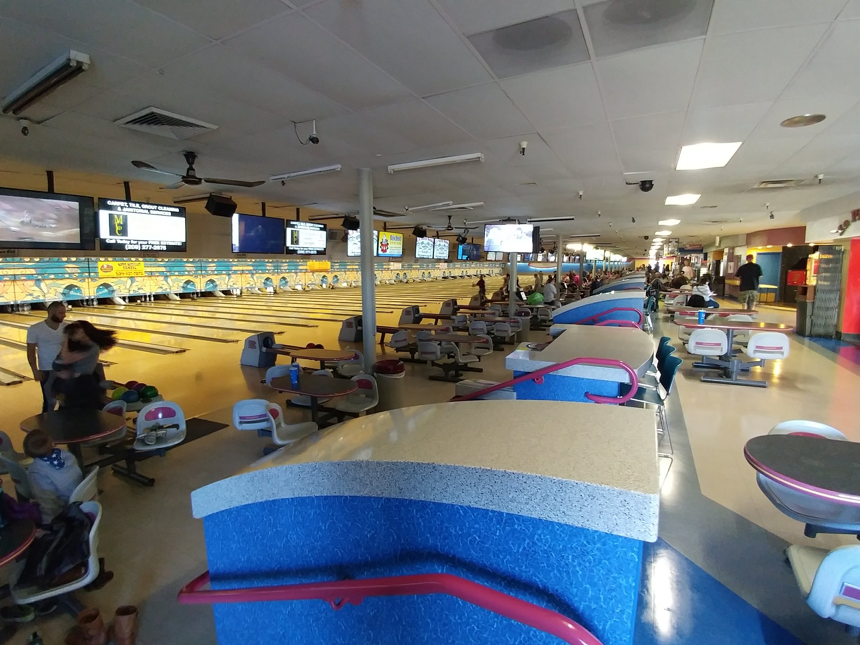 A wide interior view of a bowling alley with many lanes, overhead monitors, and blue spectator seating areas.