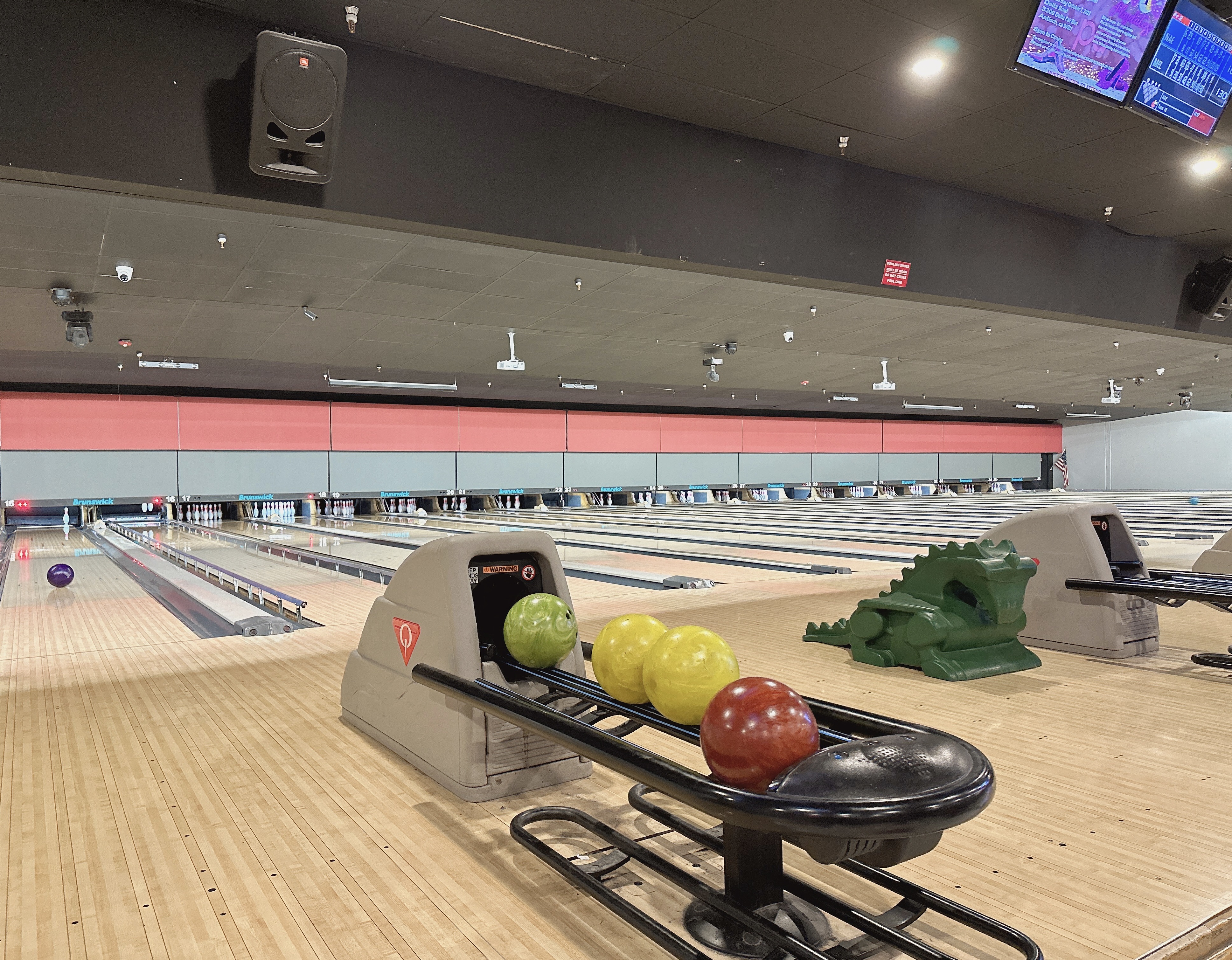 Interior view of bowling lanes with colorful balls on a return and a green dragon-shaped ramp for kids.