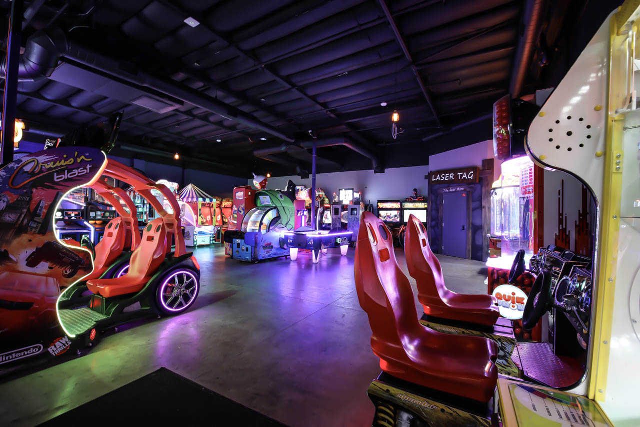 A vibrant arcade room with neon-lit racing games, an air hockey table, and a laser tag entrance.