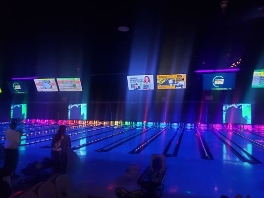 Neon blue and purple lights illuminate a row of bowling lanes during a cosmic bowling session.