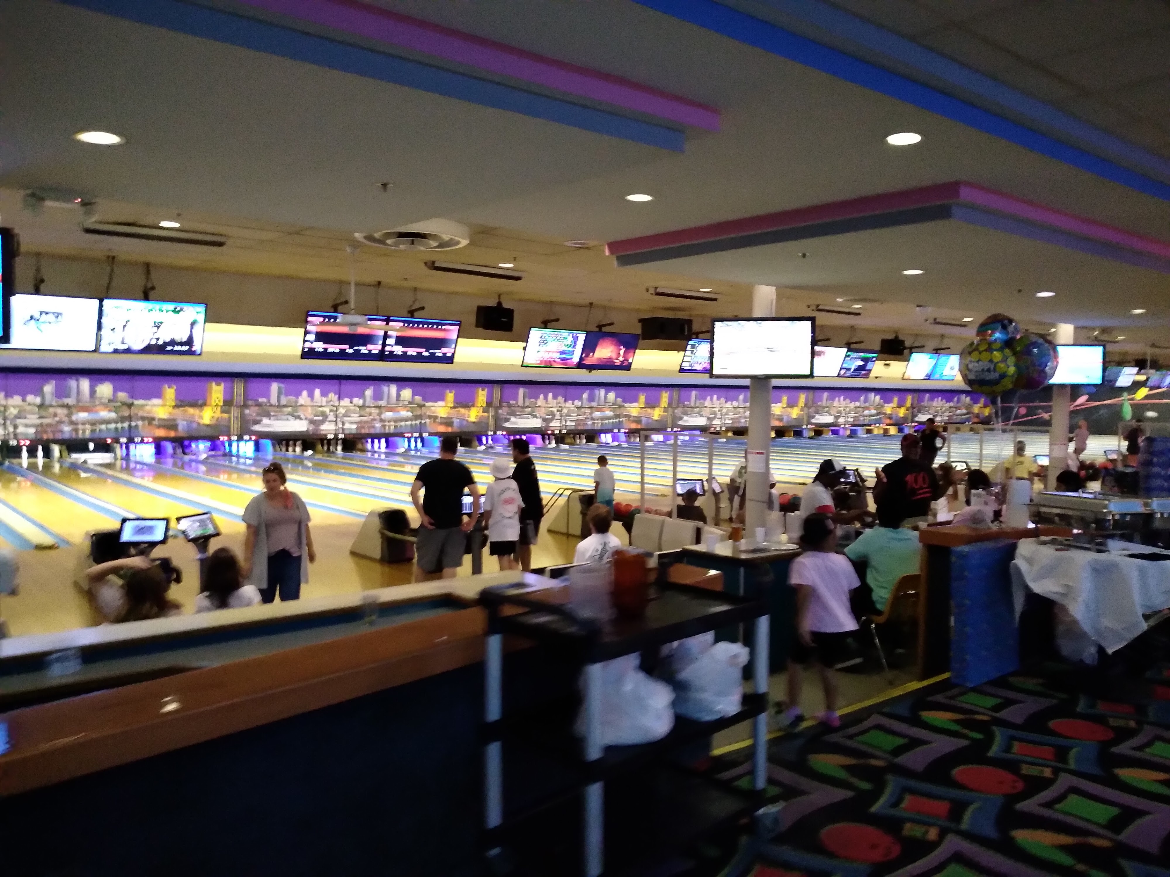 A busy bowling alley with groups playing on several lanes beneath a city skyline mural and party balloons.