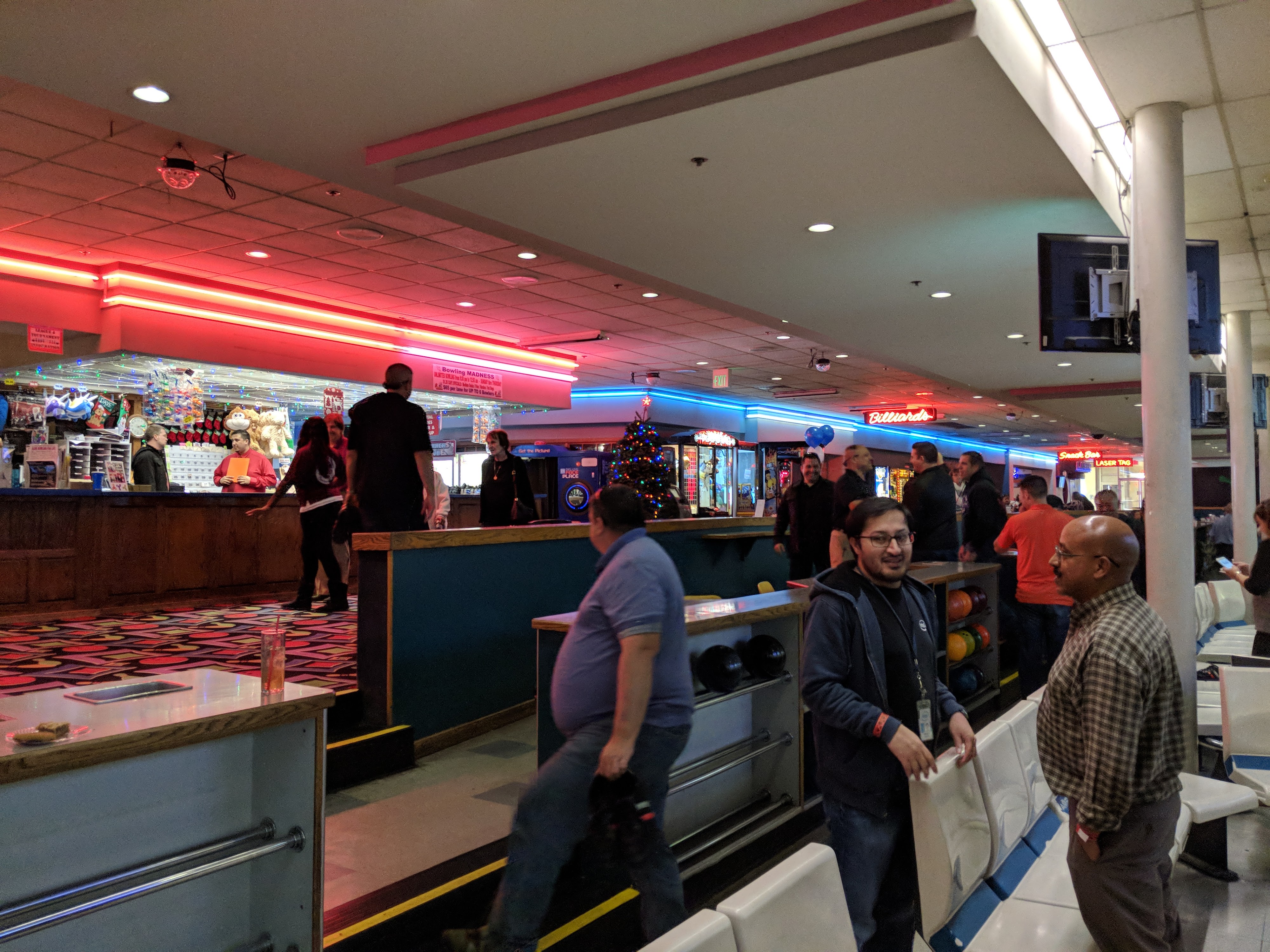Patrons socialize in the brightly lit concourse of a bowling center near a service counter and lane seating.