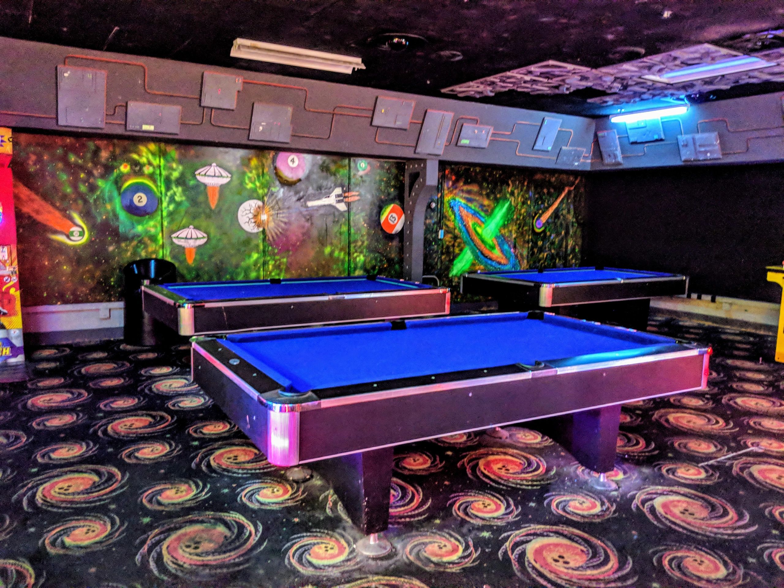 Three blue felt pool tables in a game room with space-themed murals and cosmic patterned carpet.