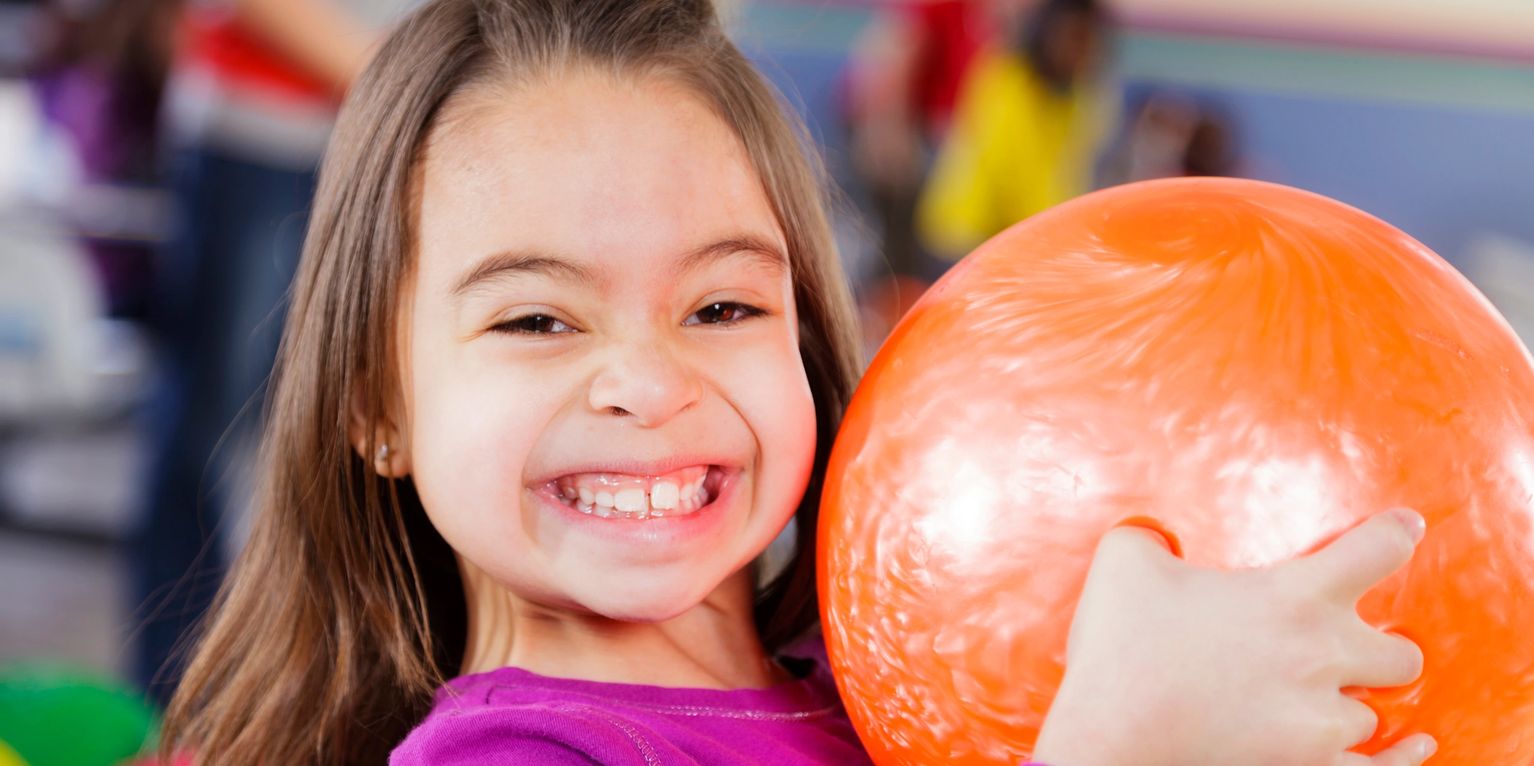 A young girl smiling broadly while holding a bright orange bowling ball.