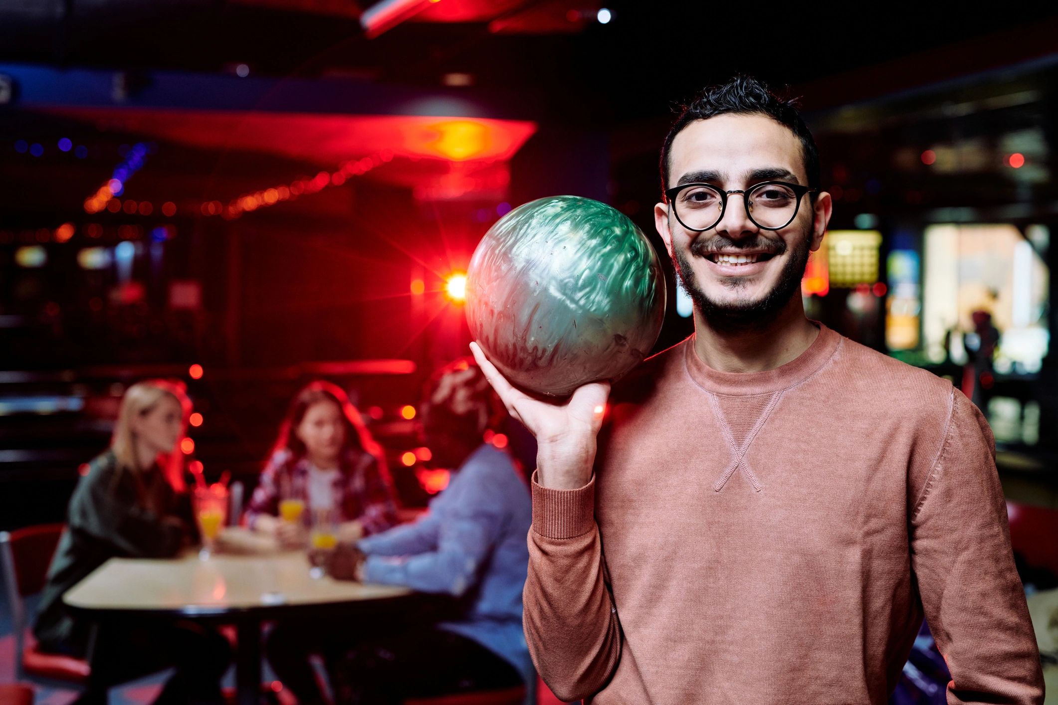 A smiling man with glasses holding a green bowling ball in a bowling center with red and blue glow lighting.