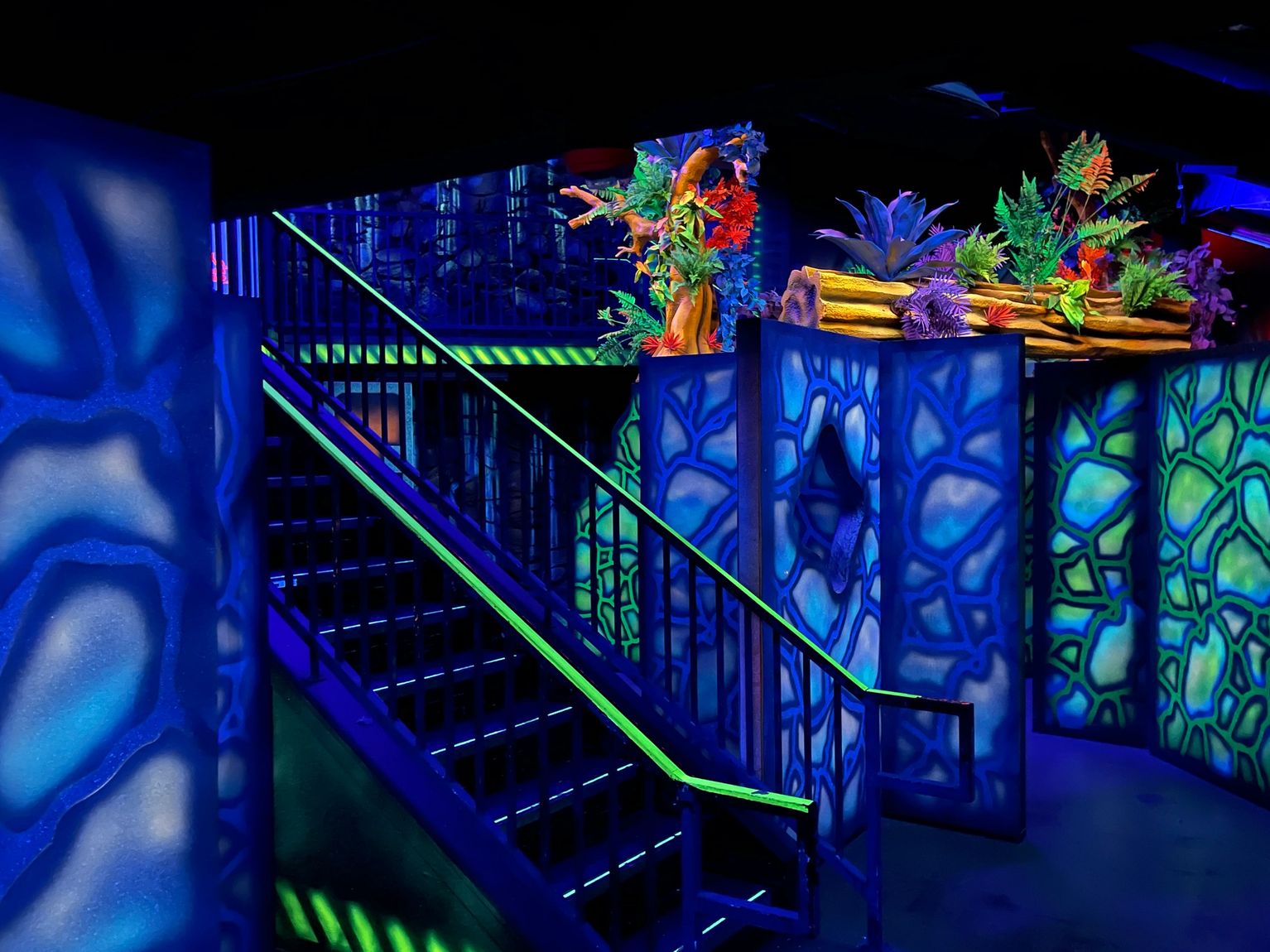 A laser tag arena with glowing neon blue and green walls and a staircase under blacklight.