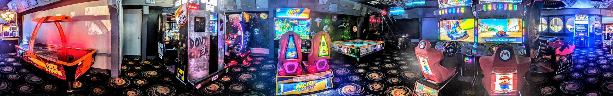 A wide panorama of an arcade room with glowing video games, air hockey, and cosmic patterned carpet.