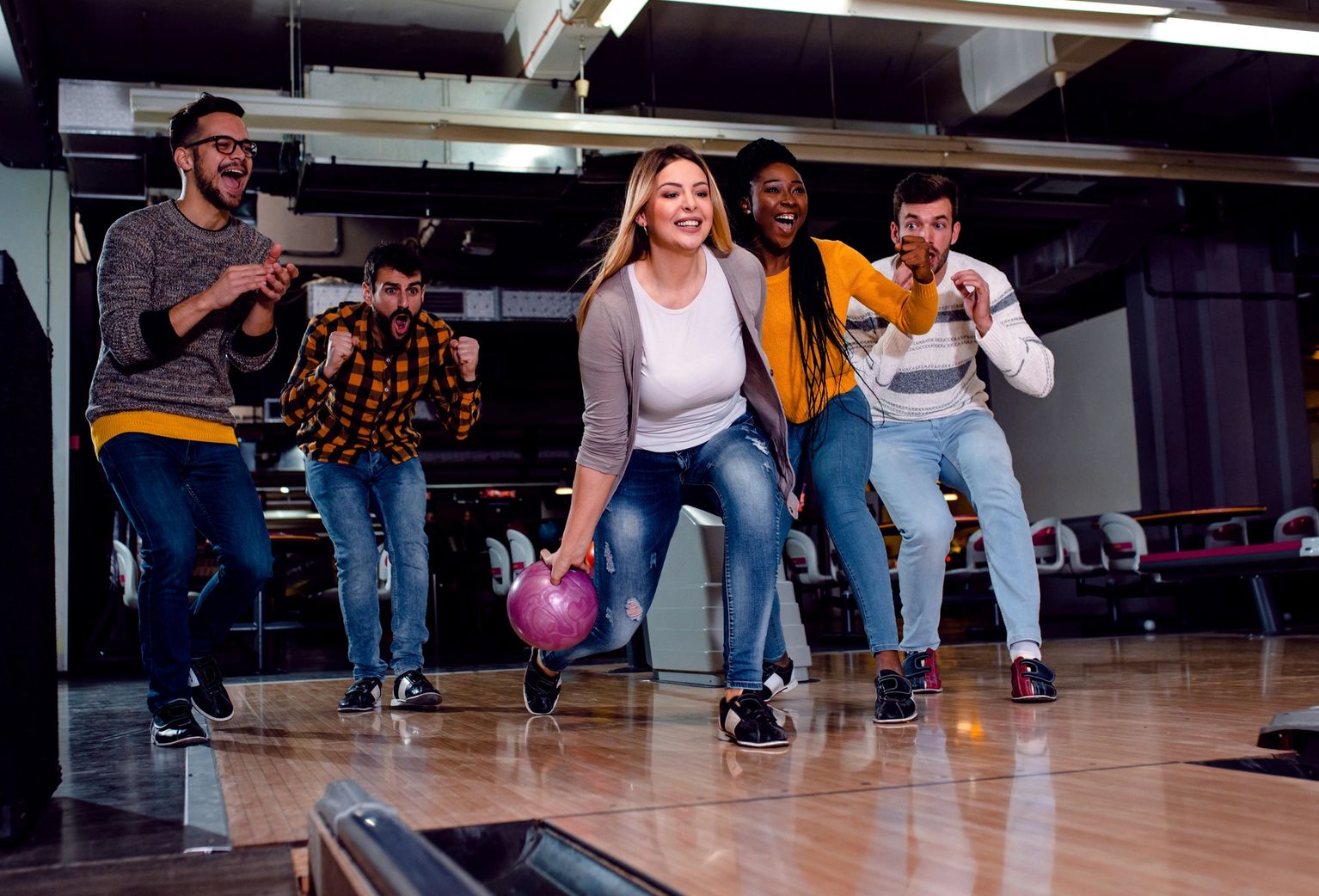 A group of friends cheers while a woman bowls a purple ball down a lane.