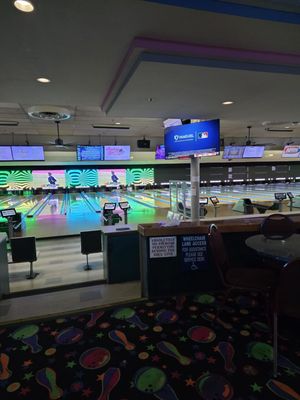 An indoor bowling alley with neon green and pink swirl graphics above the lanes and overhead television monitors.