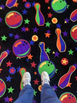 A person in white sneakers and blue jeans stands on a black carpet with neon bowling balls and pins.