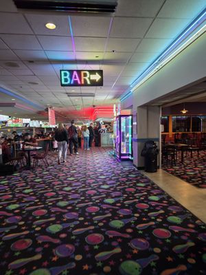 A bowling center hallway with a neon bar sign and colorful patterned carpet.