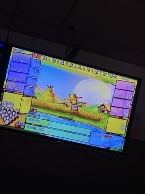 A tilted close-up of a bowling scoring monitor with cartoon farm graphics and player names.