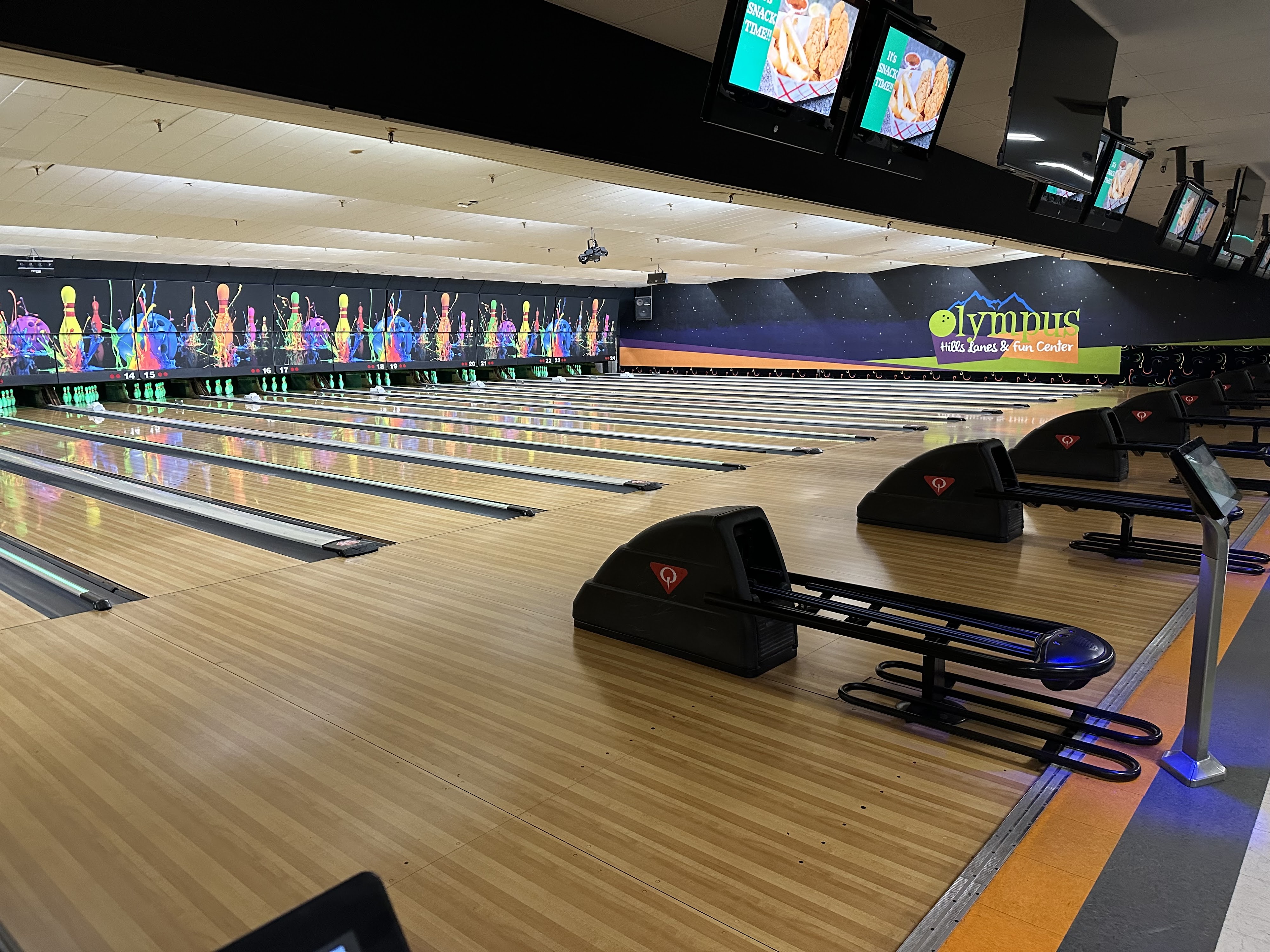 Wide view of polished wooden bowling lanes with automatic scoring monitors and a neon logo on the back wall.