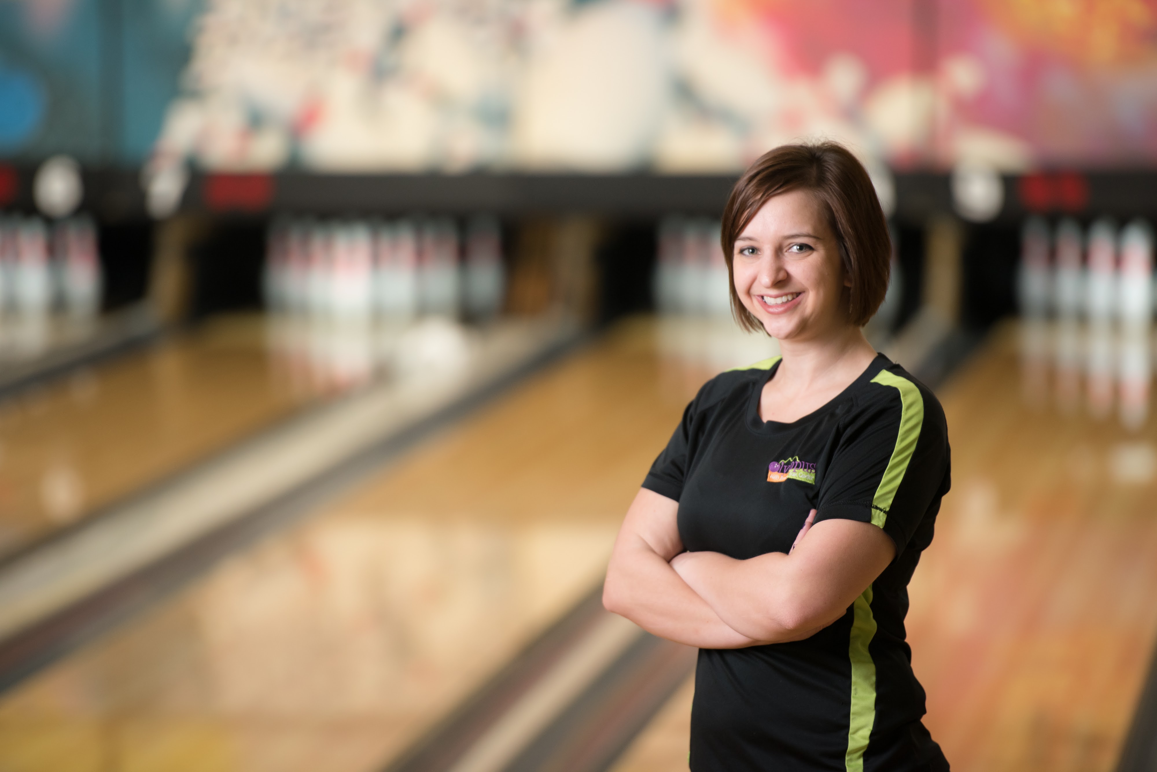 A smiling woman in a black and green shirt stands in front of blurred bowling lanes.