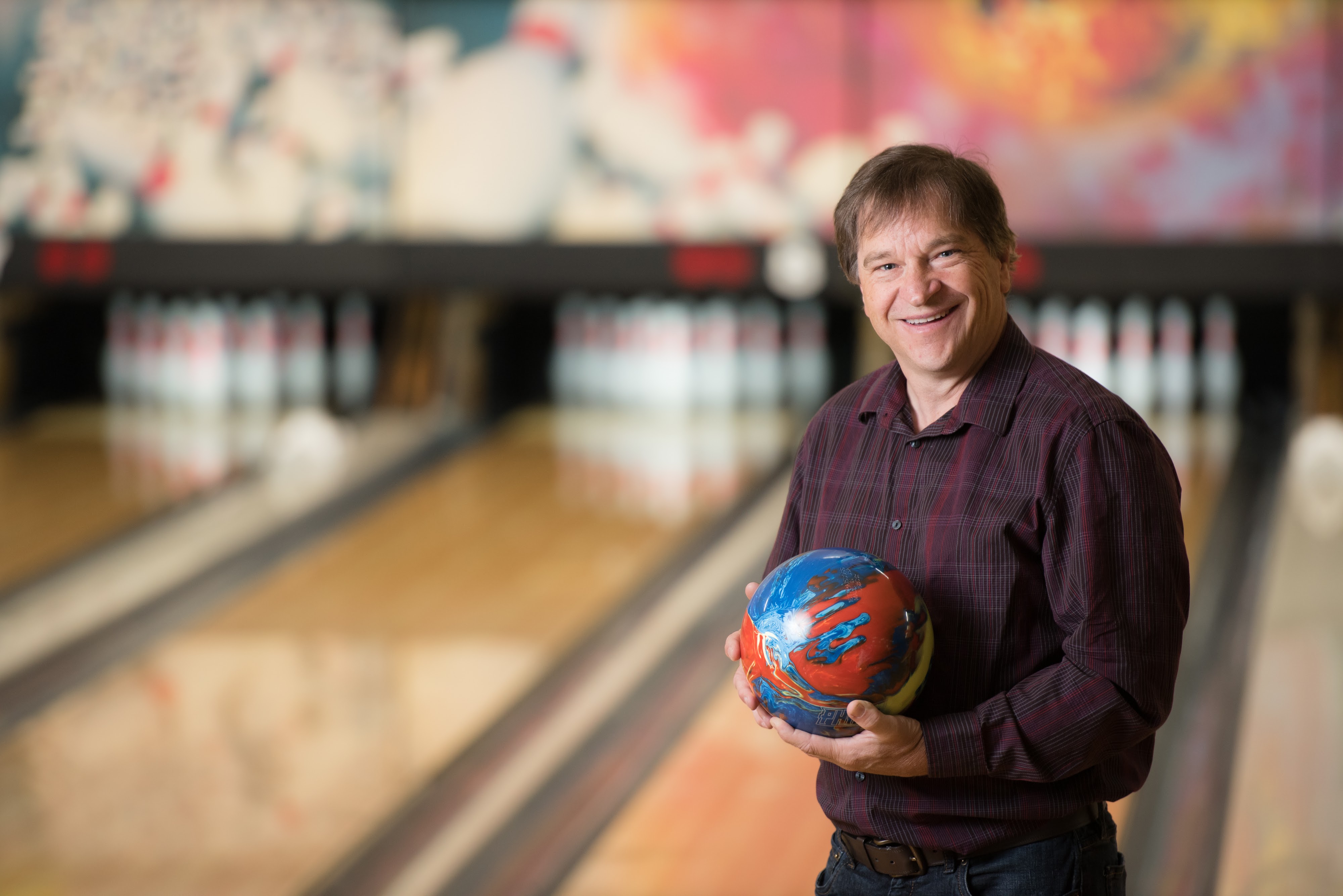 A smiling man in a maroon shirt holding a colorful bowling ball in front of blurred bowling lanes.