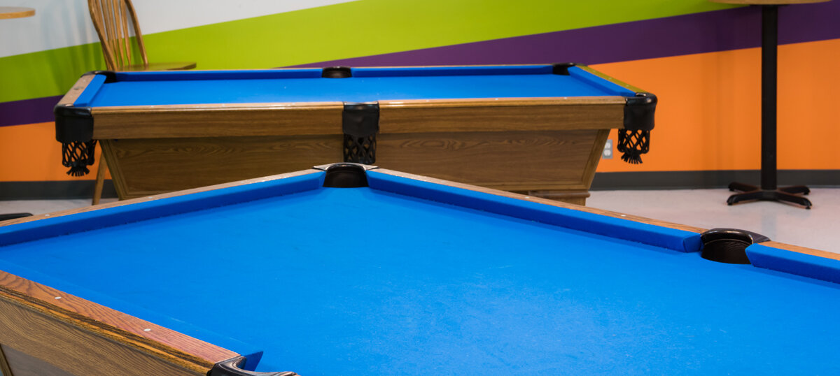 Two billiard tables with bright blue felt in a room with green, purple, and orange striped walls.