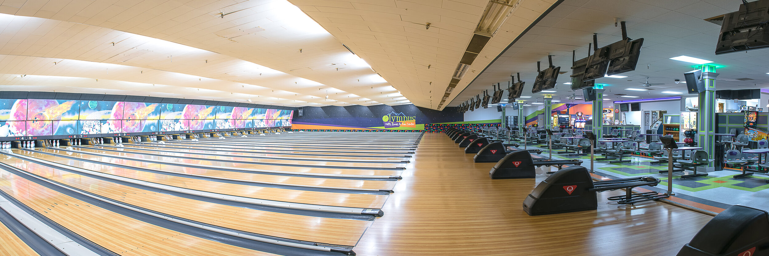 A wide panoramic view of a modern bowling center featuring multiple lanes, cosmic-themed murals, and scoring monitors.