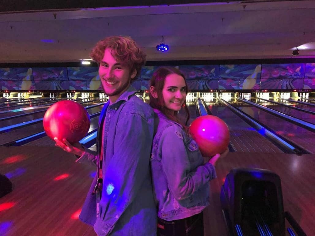 A young man and woman smiling back-to-back while holding pink bowling balls in a glow-in-the-dark bowling alley.