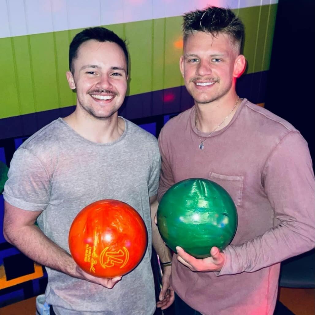 Two men smiling and holding orange and green bowling balls in a bowling alley.