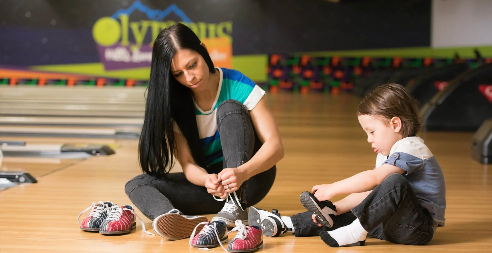 A woman and a young boy sit on a bowling lane approach putting on colorful rental bowling shoes.