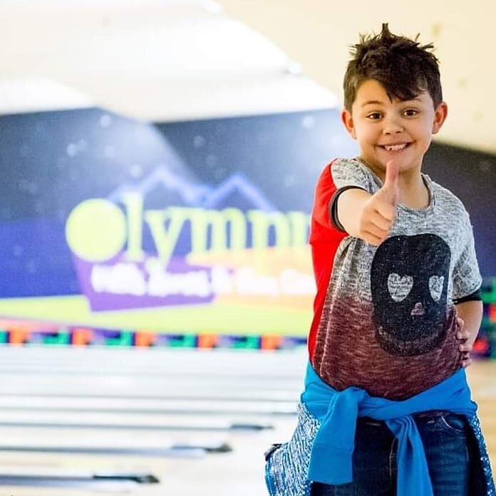 A smiling young boy giving a thumbs up in front of blurred bowling lanes and a center logo.