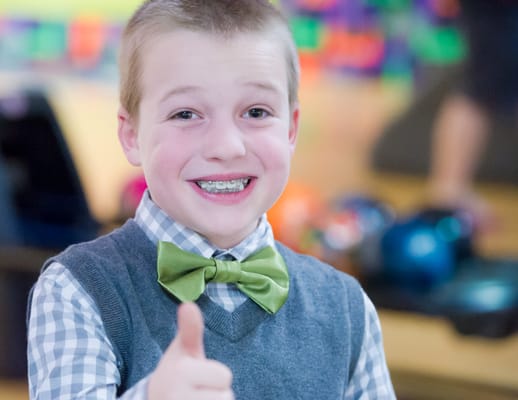 A young boy wearing a green bowtie and sweater vest gives a thumbs up at a bowling alley.